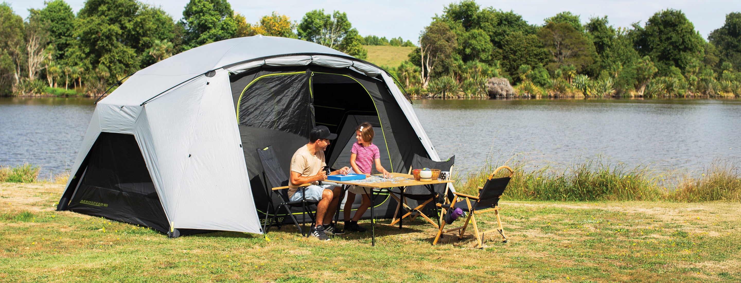 Two people sitting inside a tent by a lake with a table and chairs.