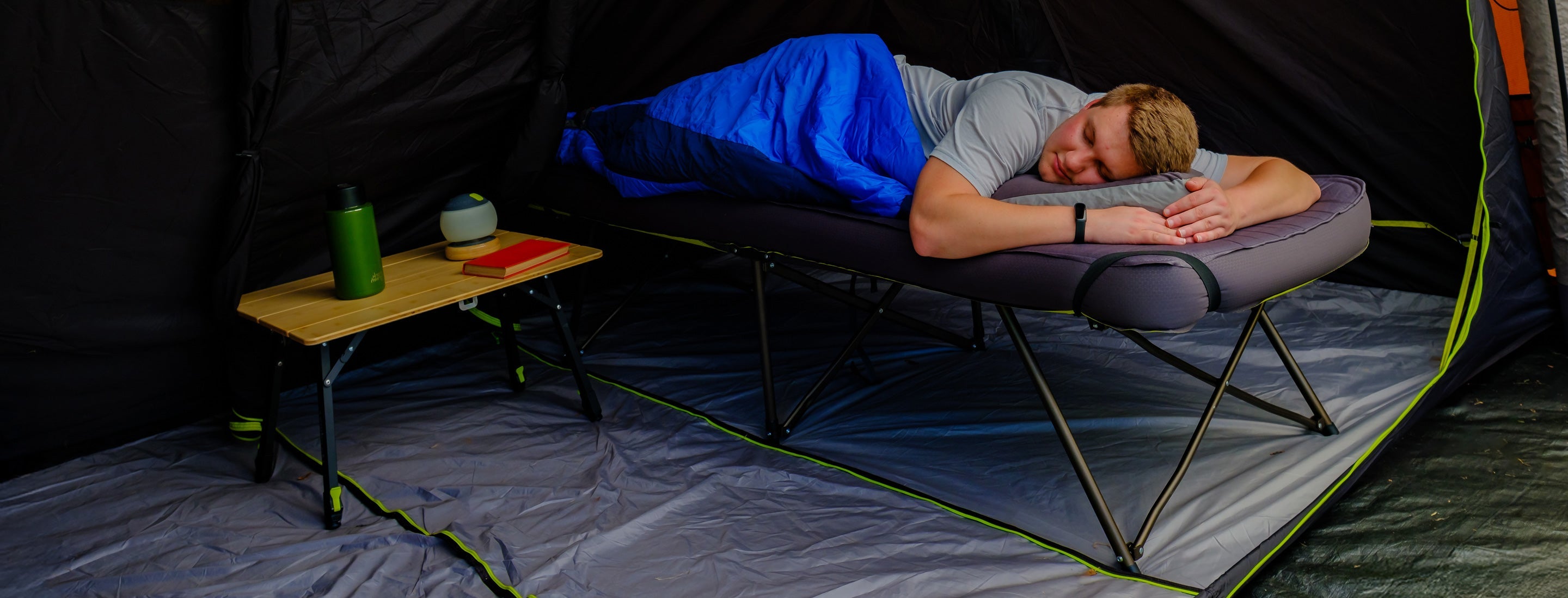 Person sleeping on a camping cot inside a tent with a small table next to them.